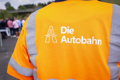 Rückenansicht einer Person, die ein reflektierendes, orangefarbenes Shirt mit der Aufschrift "Die Autobahn" trägt. Rückenansicht einer Person, die ein reflektierendes, orangefarbenes Shirt mit der Aufschrift "Die Autobahn" trägt.