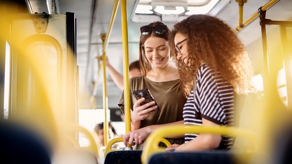 Zum Artikel Zwei junge Frauen stehen in einem Bus, schauen auf ein Handy und lächeln